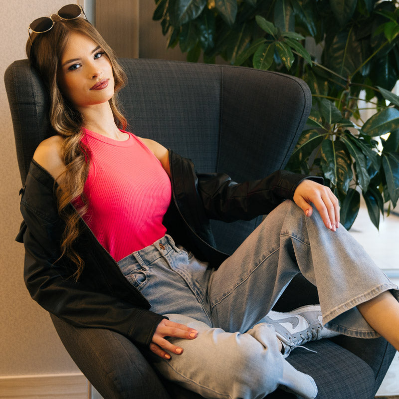 Model sitting on chair wearing hot pink tank, wide leg light wash jeans, biker faux leather jacket, and sunglasses.