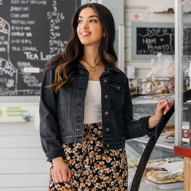Model smiling standing in a cafe, wearing a black denim jacket over a white shirt and floral skirt.