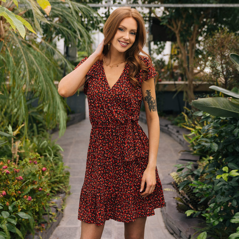 Smiling model standing in greenhouse wearing a red and orange-toned floral crossover skater dress.