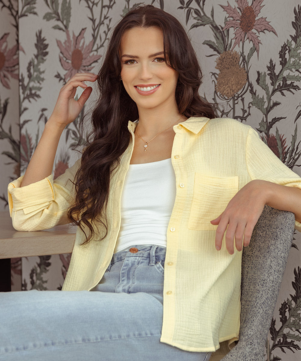 Model smiling and looking at the camera, wearing a pale yellow cotton button-up shirt over a white tank top and light wash jeans.